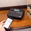 Brother P-touch label maker with keyboard and display on desk with hand holding smartphone