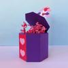 Purple hexagonal gift box with red heart panel, open lid, and pink paper confetti bursting out