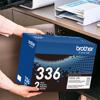 Person holding Brother TN336 2-pack toner cartridge box in home office with printer