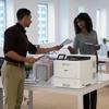 Two professionals sharing documents beside Brother color laser printer in modern office