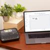 Brother label maker on wooden desk with laptop displaying label design software and plants