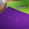 Brother P-touch label on green notebook beside purple notebook with spiral binding